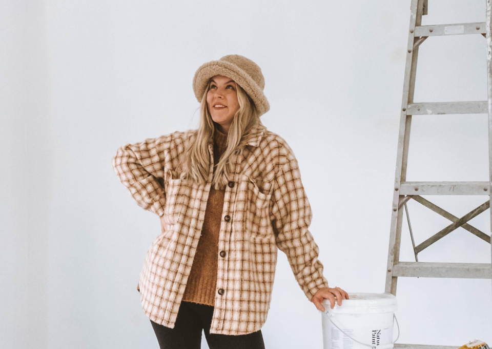 Woman in plaid shirt and hat standing next to a ladder against a white wall