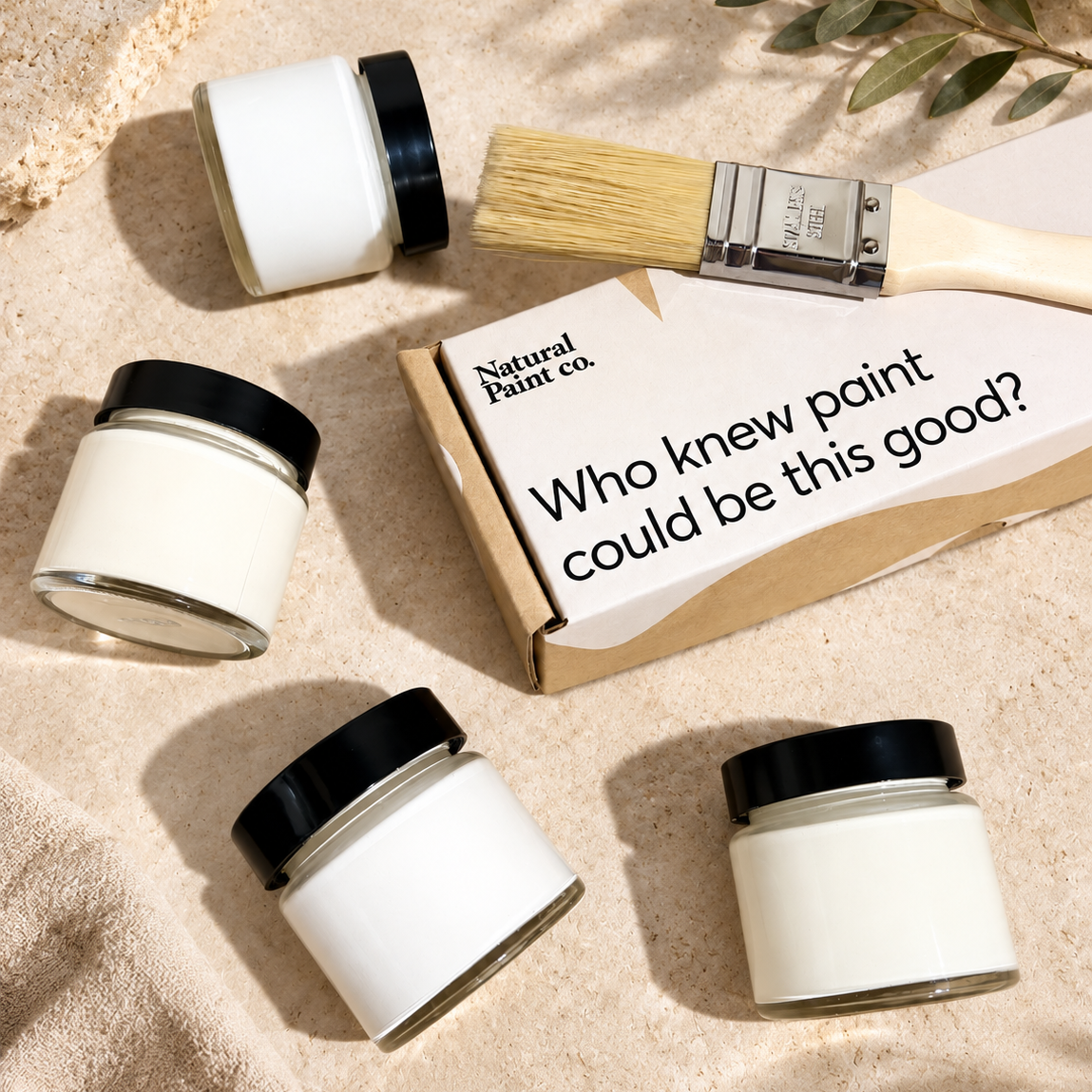 Four white paint jars with black lids are arranged on a beige textured surface with a paintbrush and a Natural Paint Co. box.